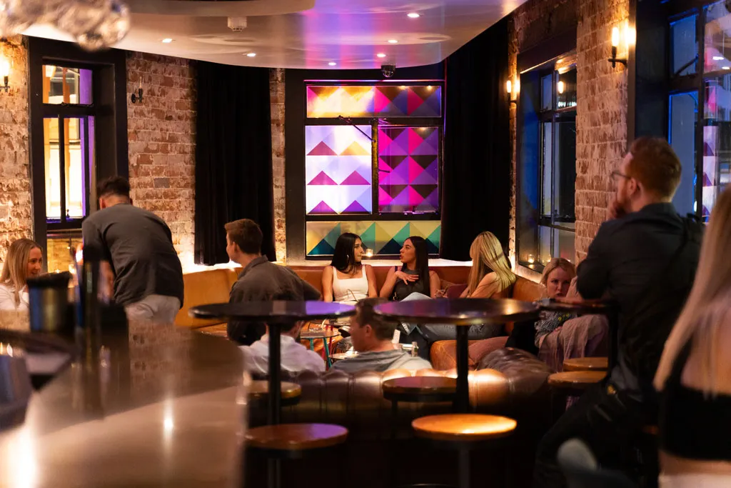 Groups of friends at low tables in the Humber cocktail lounge, with exposed brick walls, a brass bar and a coloured glass window