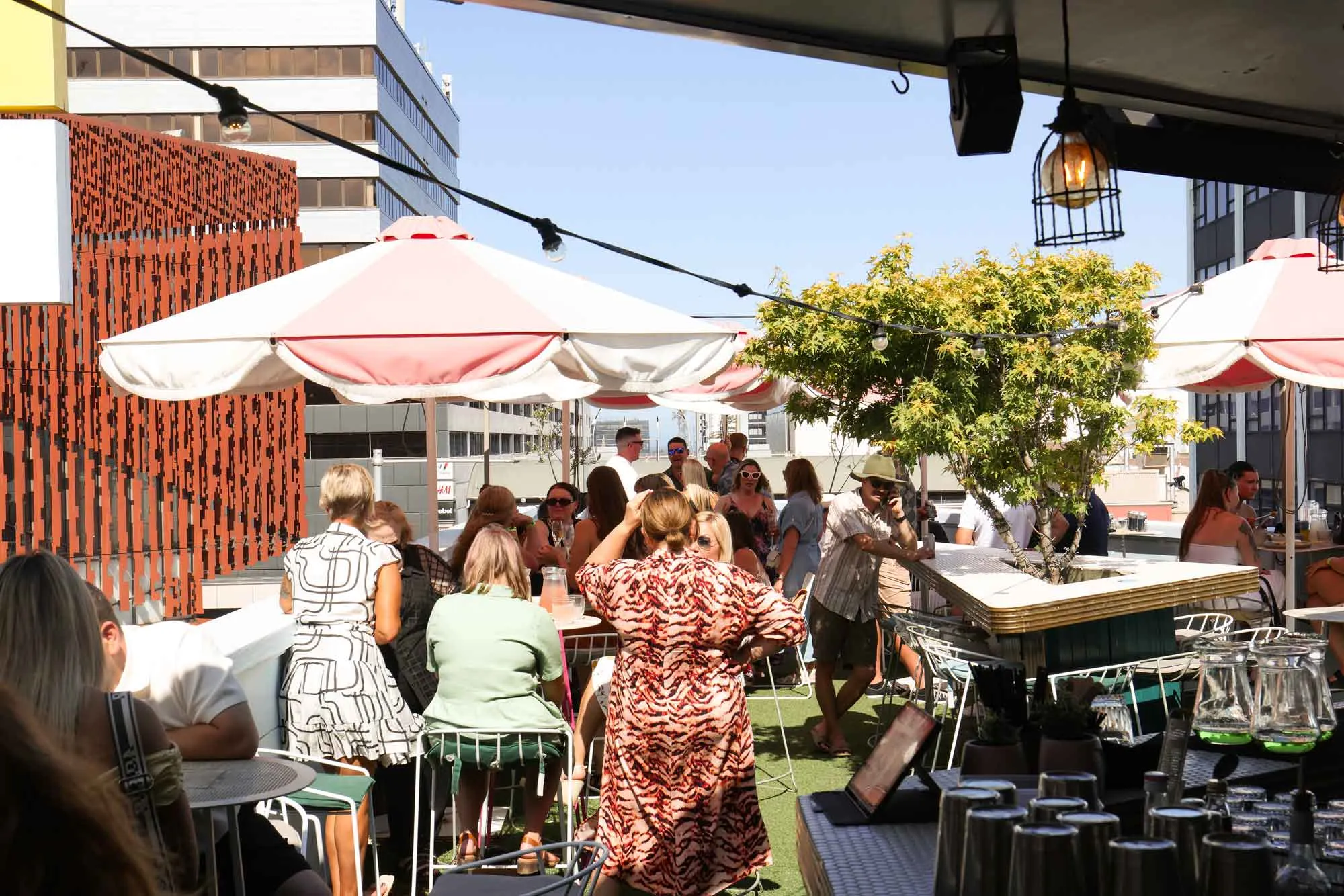 A lively crowd drinking under pink and white umbrellas on the Humber rooftop, with the brass bar and city skyline behind